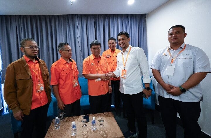 Handshake between a man in a white shirt and a man in an orange shirt, with four colleagues watching in a small conference room.