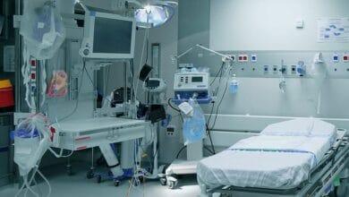 Empty hospital bed in a sterile ICU with a wall of monitors, IV lines, and life-support equipment nearby.