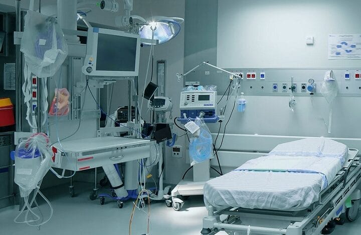 Empty hospital bed in a sterile ICU with a wall of monitors, IV lines, and life-support equipment nearby.