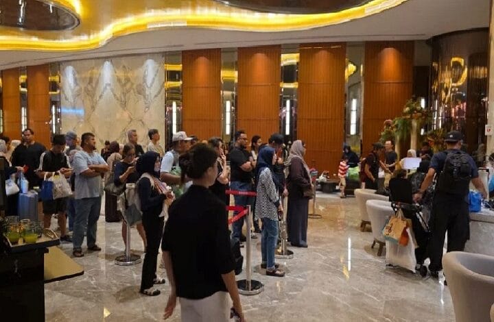 People standing in a queue in a hotel lobby with polished floors, wood paneled walls, and warm yellow lighting.
