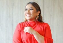 Woman in a red blouse smiles while looking upward, hands gently resting at chest, indoors with light wood background