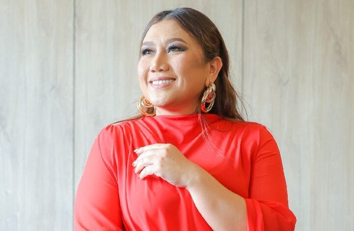 Woman in a red blouse smiles while looking upward, hands gently resting at chest, indoors with light wood background