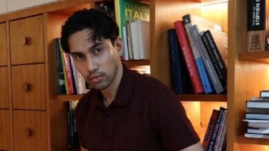 idris khan 2 - Wilayah.com.my Man in a brown polo shirt posing in front of a wooden bookshelf filled with cookbooks in a cozy library setting. Readable book spines are visible behind him.