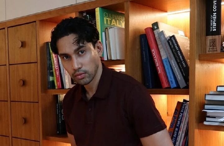 idris khan 2 - Wilayah.com.my Man in a brown polo shirt posing in front of a wooden bookshelf filled with cookbooks in a cozy library setting. Readable book spines are visible behind him.