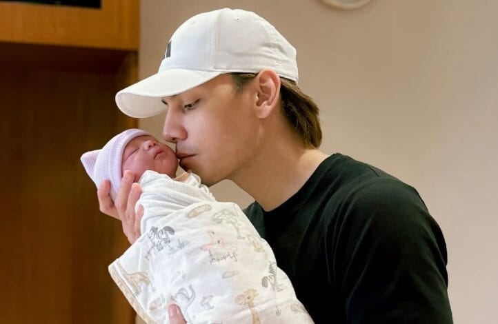 A man in a white cap gently cradles and kisses a newborn wrapped in a patterned blanket.