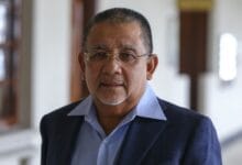 Portrait of a middle-aged man with short gray hair and glasses, wearing a blue shirt and navy blazer in a hallway at an indoor setting.
