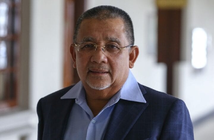 Portrait of a middle-aged man with short gray hair and glasses, wearing a blue shirt and navy blazer in a hallway at an indoor setting.