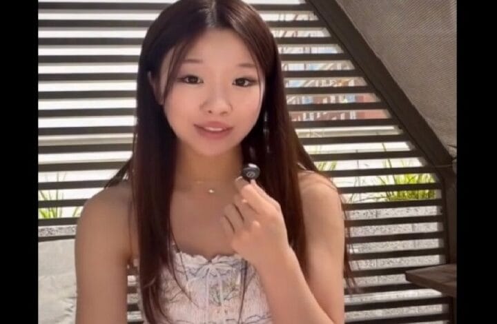 jaime tan - Wilayah.com.my Young woman with long dark hair in a white patterned sleeveless top holds a small lapel microphone toward the camera.