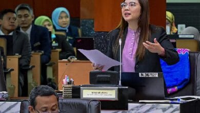 jamilah 2 - Wilayah.com.my A woman in a blazer speaking at a podium in a meeting room, holding papers and gesturing as she presents to an audience seated behind her.