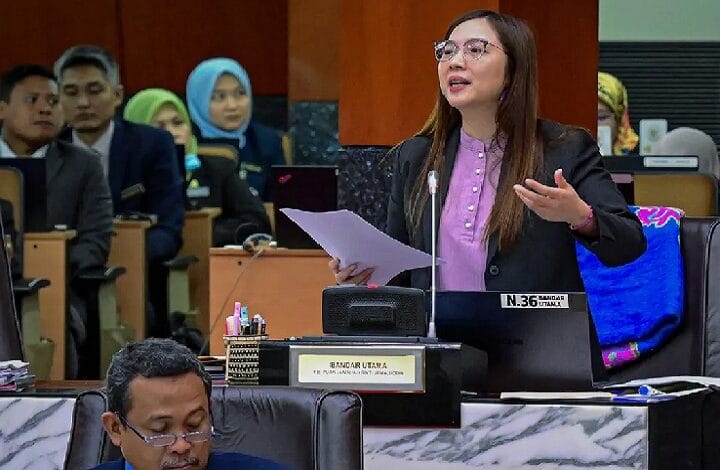 jamilah 2 - Wilayah.com.my A woman in a blazer speaking at a podium in a meeting room, holding papers and gesturing as she presents to an audience seated behind her.