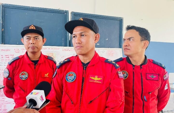Three men in red flight suits stand indoors, one speaking into a microphone during a press briefing.