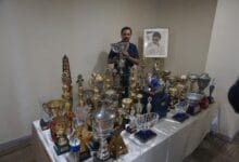karamjit singh 2 - Wilayah.com.my Man standing behind a table covered with many trophies and medals, with a framed portrait on the wall behind him.