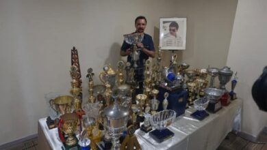 karamjit singh 2 - Wilayah.com.my Man standing behind a table covered with many trophies and medals, with a framed portrait on the wall behind him.