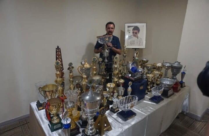 karamjit singh 2 - Wilayah.com.my Man standing behind a table covered with many trophies and medals, with a framed portrait on the wall behind him.