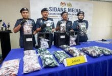 Four police officers in uniform display bags of seized drugs on a blue-table at a press conference, with a 'DADAH' sign in front.