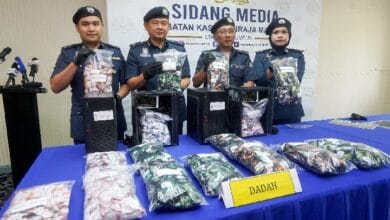 Four police officers in uniform display bags of seized drugs on a blue-table at a press conference, with a 'DADAH' sign in front.