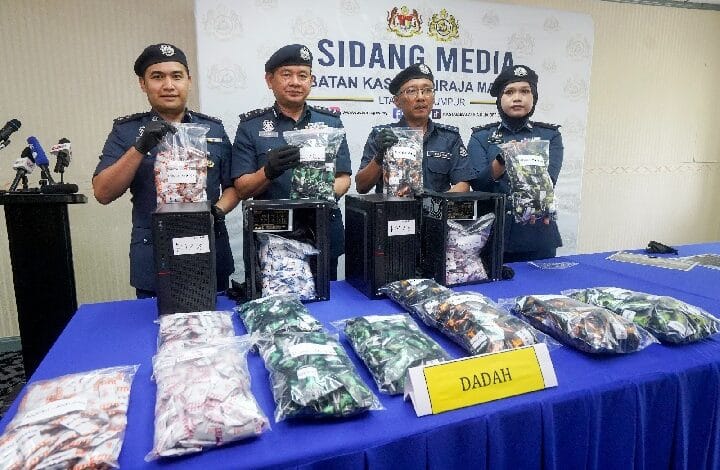 Four police officers in uniform display bags of seized drugs on a blue-table at a press conference, with a 'DADAH' sign in front.