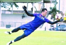 khairul apek - Wilayah.com.my Soccer goalkeeper in a blue kit dives mid-air to reach the ball on a grassy field during a match as gloves flap
