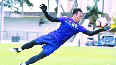 Soccer goalkeeper in a blue kit dives mid-air to reach the ball on a grassy field during a match as gloves flap