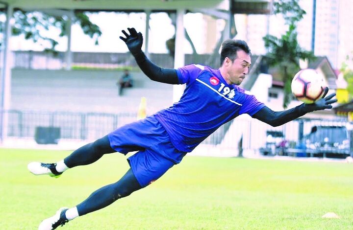 Soccer goalkeeper in a blue kit dives mid-air to reach the ball on a grassy field during a match as gloves flap