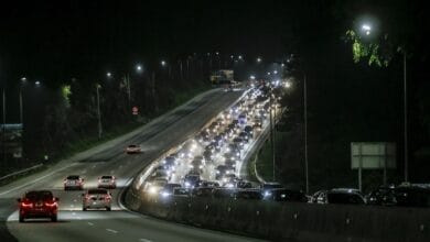Nighttime highway with heavy traffic and bright headlights on both directions, streetlights lining the road.