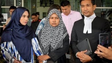 lab assistant 2 - Wilayah.com.my Three people stand outside a glass-front building: two women in hijabs and a man in a black suit with a white neckband, as a bystander films with a phone.