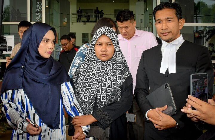 lab assistant 2 - Wilayah.com.my Three people stand outside a glass-front building: two women in hijabs and a man in a black suit with a white neckband, as a bystander films with a phone.