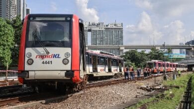 lrt ampang - Wilayah.com.my Red and white RapidKL train on tracks with maintenance workers in orange vests nearby and city buildings in the background.