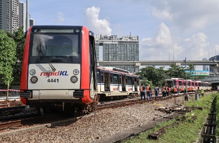 Red and white RapidKL train on tracks with maintenance workers in orange vests nearby and city buildings in the background.