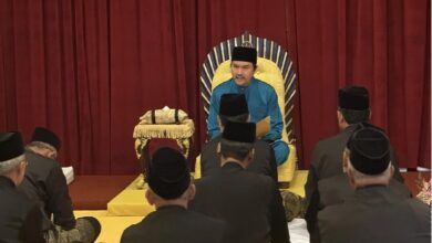 A man in blue traditional attire sits on a gold throne on stage, addressing an audience of men in black suits and caps.