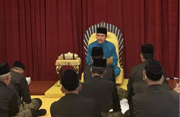 A man in blue traditional attire sits on a gold throne on stage, addressing an audience of men in black suits and caps.