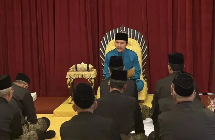A man in blue traditional attire sits on a gold throne on stage, addressing an audience of men in black suits and caps.