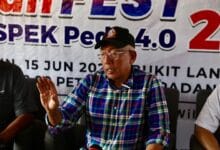 mahdzir - Wilayah.com.my Older man wearing a blue-green plaid shirt and black cap speaks at a press conference, gesturing with his left hand; banner behind shows event details.