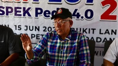 Older man wearing a blue-green plaid shirt and black cap speaks at a press conference, gesturing with his left hand; banner behind shows event details.