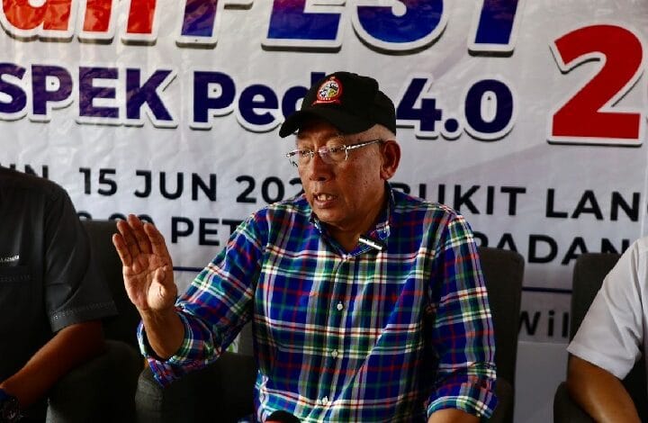 Older man wearing a blue-green plaid shirt and black cap speaks at a press conference, gesturing with his left hand; banner behind shows event details.