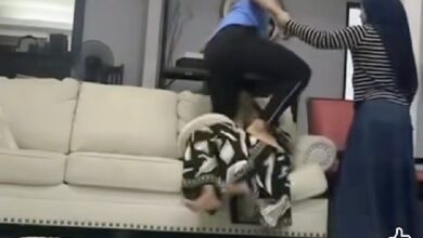 maid dera - Wilayah.com.my Two people in a living room struggle: one person on a sofa is grabbed by another standing over them, while a third person in a striped shirt watches nearby.