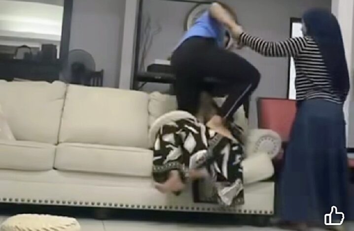 Two people in a living room struggle: one person on a sofa is grabbed by another standing over them, while a third person in a striped shirt watches nearby.