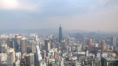 malaysia - Wilayah.com.my Wide cityscape of a dense modern skyline with a tall dark skyscraper in the center under a hazy sky.