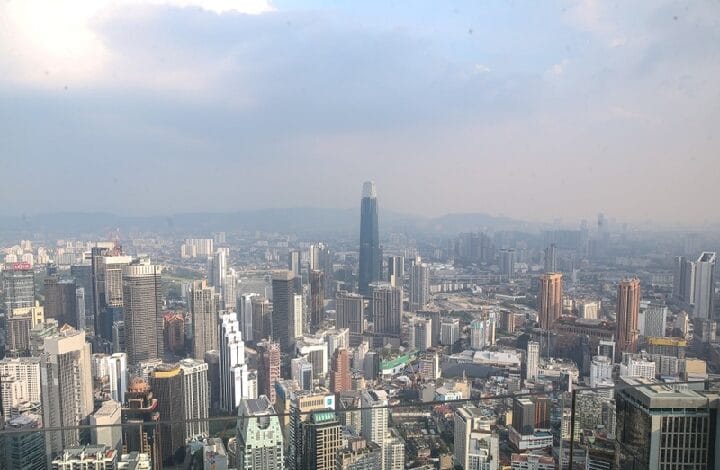 malaysia - Wilayah.com.my Wide cityscape of a dense modern skyline with a tall dark skyscraper in the center under a hazy sky.