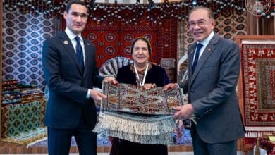 malaysia turkey - Wilayah.com.my Three people in formal attire present a folded decorative rug in a room filled with colorful carpets and patterns.