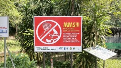 Red no-smoking warning sign in Indonesian with a crossed-out cigarette symbol, posted in a garden setting.