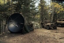 Soldiers in a forest clearing deploy a large satellite dish next to armored vehicles during a field operation.
