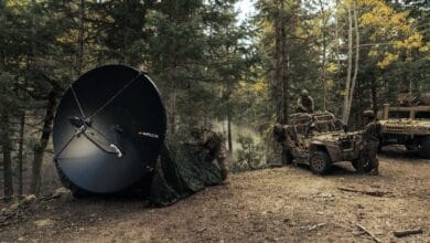 Soldiers in a forest clearing deploy a large satellite dish next to armored vehicles during a field operation.