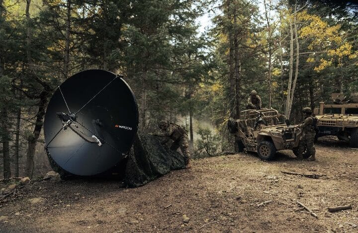 Soldiers in a forest clearing deploy a large satellite dish next to armored vehicles during a field operation.