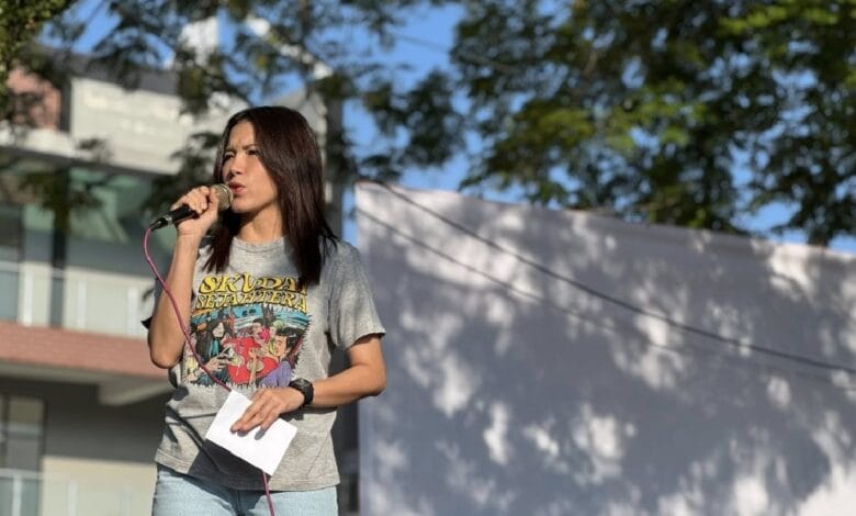 Woman singing into a microphone on an outdoor stage, holding a small sheet of paper, wearing a gray graphic T-shirt.