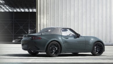 Teal Mazda MX-5 Miata convertible with a white roof, parked near a metal industrial building.