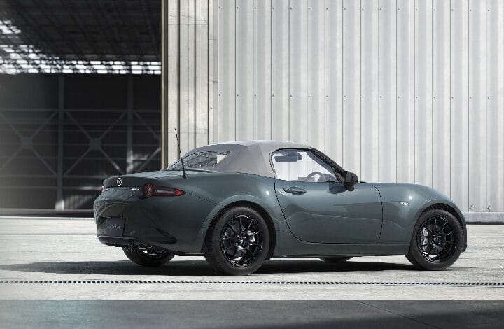 Teal Mazda MX-5 Miata convertible with a white roof, parked near a metal industrial building.
