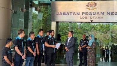 Group of men in matching polo shirts listen to a man in a suit outside the main lobby of Jabatan Peguam Negara (Attorney General's Office).