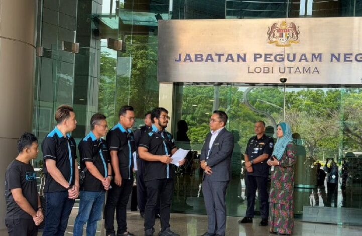 Group of men in matching polo shirts listen to a man in a suit outside the main lobby of Jabatan Peguam Negara (Attorney General's Office).