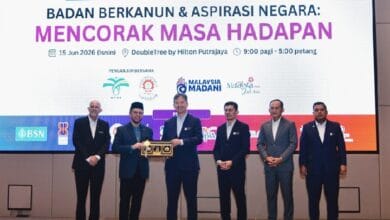 mof kewangan - Wilayah.com.my Six men in suits stand on a stage as one hands a wooden trophy to another, with a large screen behind them displaying an event banner in Malay.